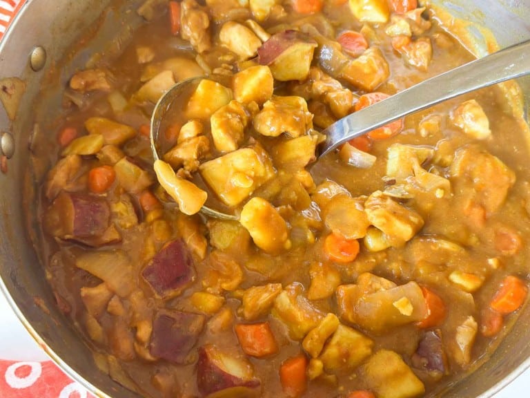 A ladle scoops chicken curry with potatoes, carrots, and onions from a metal pot.