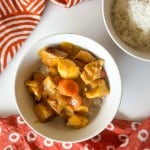 A bowl of chicken curry with potatoes and carrots served over white rice, alongside a separate bowl of plain rice, on a white surface with red patterned cloths.