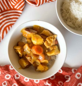 A bowl of chicken curry with potatoes and carrots served over white rice, alongside a separate bowl of plain rice, on a white surface with red patterned cloths.