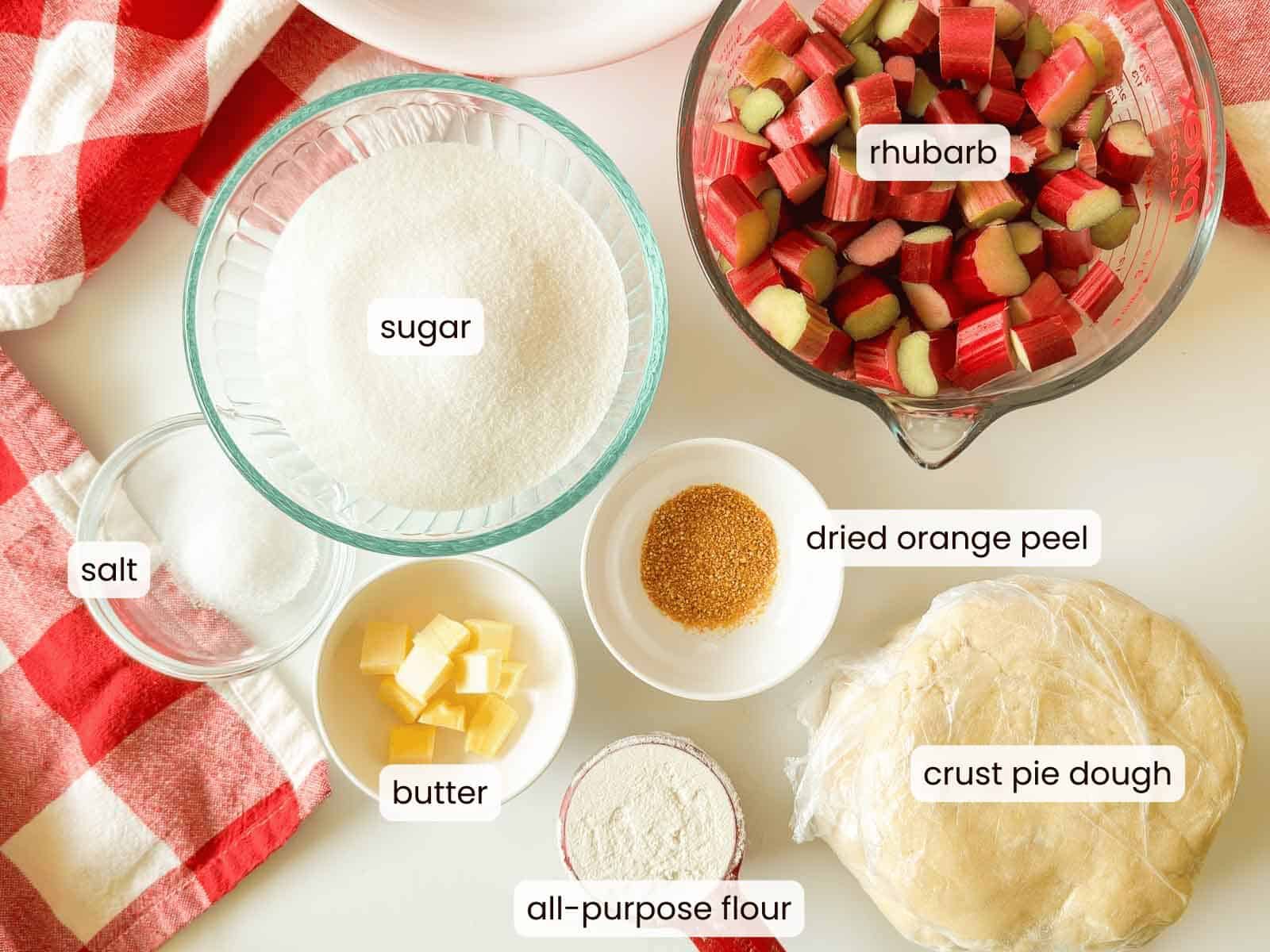 Top-down view of labeled ingredients for classic rhubarb pie, including rhubarb, sugar, dried orange peel, salt, butter, all-purpose flour, and crust pie dough arranged on a countertop.