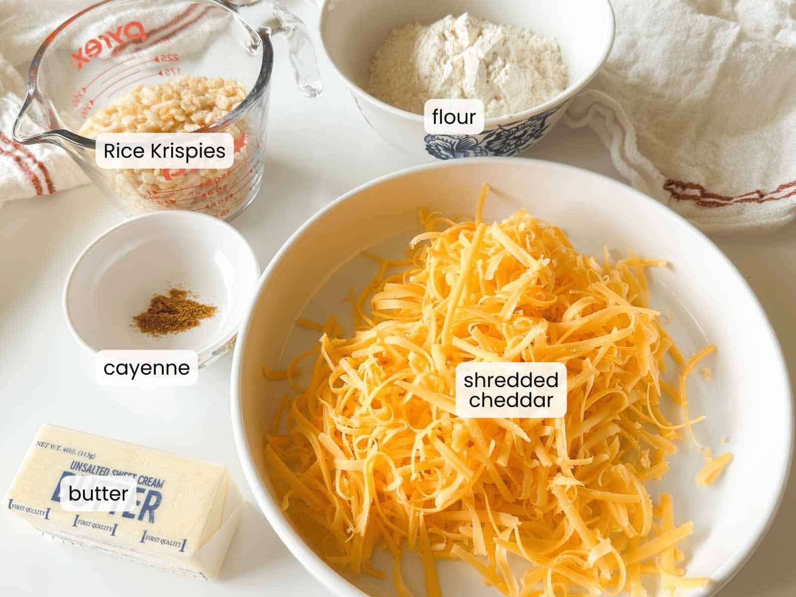 A bowl of shredded cheddar, a stick of butter, a small bowl of cayenne, a measuring cup of Rice Krispies, and a bowl of flour are arranged on a white surface—perfect ingredients for making homemade Rice Krispie Crackers.