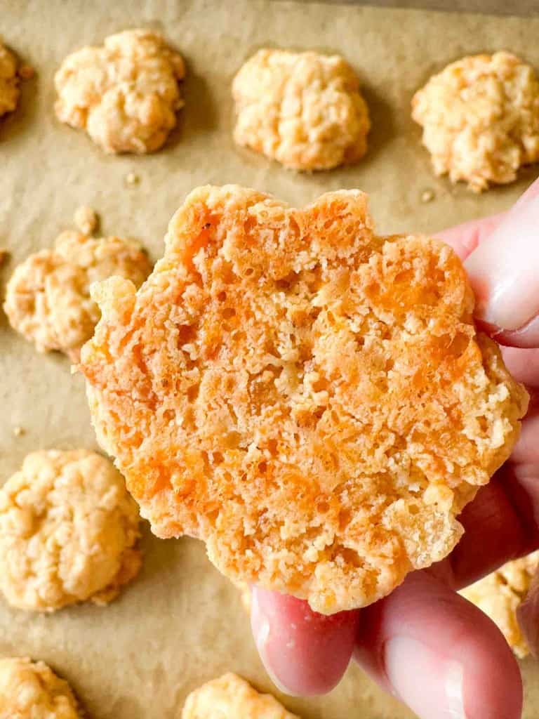 A hand holds up a cheesy, golden-brown biscuit with a crumbly texture; more biscuits are visible on a parchment-lined baking tray in the background.