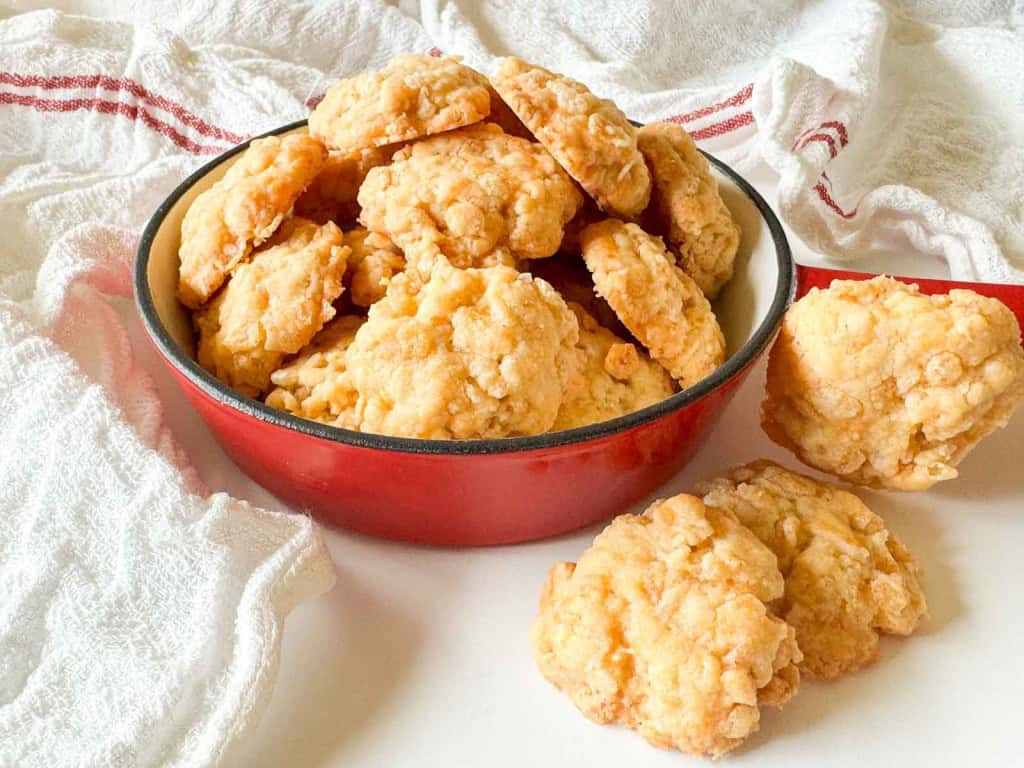 A red bowl filled with golden, crumbly cheese biscuits sits on a white surface, with a few biscuits resting nearby and a white towel in the background.