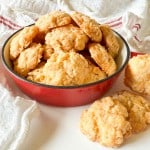 A red bowl filled with golden, crumbly cheese biscuits sits on a white surface, with a few biscuits resting nearby and a white towel in the background.