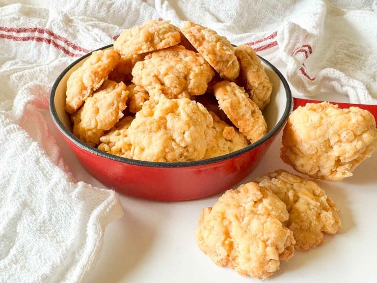 A red bowl filled with golden, crumbly cheese biscuits sits on a white surface, with a few biscuits resting nearby and a white towel in the background.