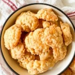A white bowl filled with a pile of golden, crumbly cheese biscuits sits on a white surface with a textured cloth in the background.