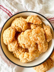 A white bowl filled with a pile of golden, crumbly cheese biscuits sits on a white surface with a textured cloth in the background.
