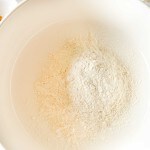 A white mixing bowl containing a mound of flour on a white surface, with the edge of a measuring cup and another bowl partially visible.
