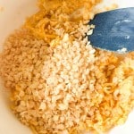 A close-up of a mixing bowl containing shredded cheese and crispy rice cereal being stirred with a blue spatula.