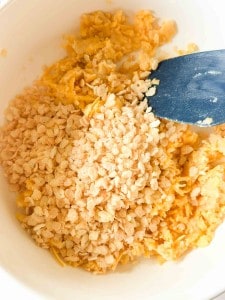 A close-up of a mixing bowl containing shredded cheese and crispy rice cereal being stirred with a blue spatula.
