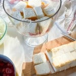 Cubed pieces of white cake are layered in a glass trifle bowl. More cubes sit on a wooden cutting board nearby, next to a knife and bowls with ingredients.