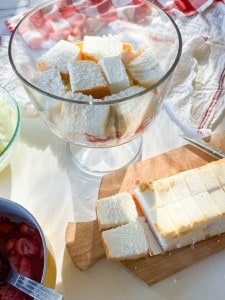 Cubed pieces of white cake are layered in a glass trifle bowl. More cubes sit on a wooden cutting board nearby, next to a knife and bowls with ingredients.