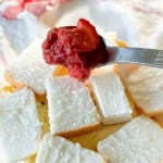 A fork holds strawberry topping above a bowl of cubed white cake pieces.