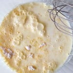 A mixing bowl containing a bubbly liquid batter with visible lumps and a metal whisk resting on the side.
