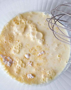 A mixing bowl containing a bubbly liquid batter with visible lumps and a metal whisk resting on the side.