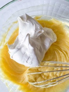 A whisk rests in a bowl of yellow batter topped with a large dollop of whipped cream or meringue.