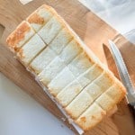 A loaf of cake sliced into twelve square pieces sits on a wooden cutting board next to a serrated knife.