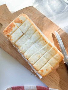 A loaf of cake sliced into twelve square pieces sits on a wooden cutting board next to a serrated knife.