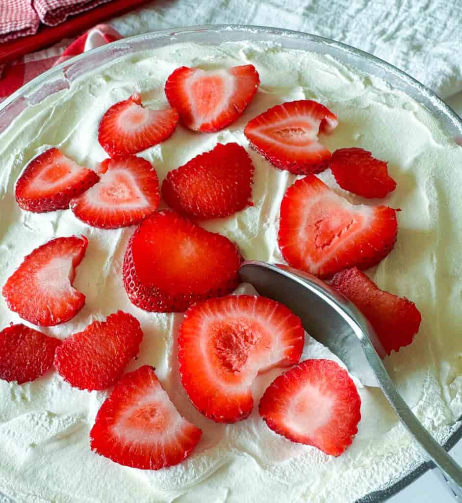 A glass dish filled with whipped cream, topped with sliced strawberries, evokes a classic Strawberry Rhubarb Trifle, with a metal spoon resting on the surface ready for a delicious bite.