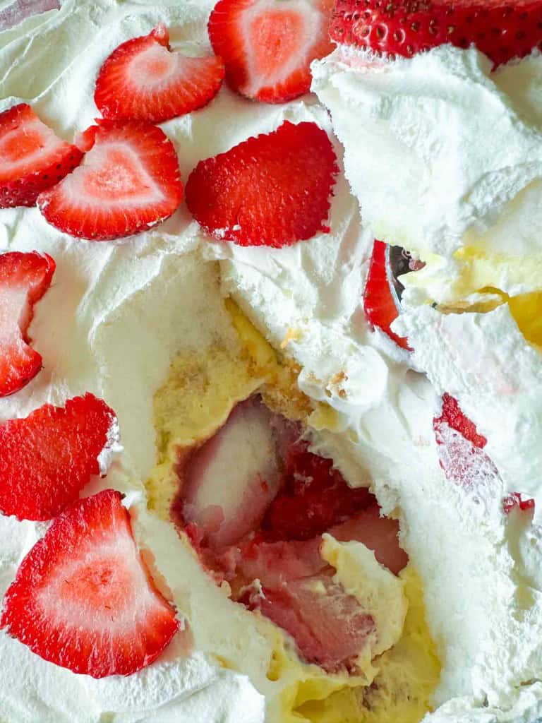 Close-up of a Strawberry Rhubarb Trifle with whipped cream, sliced strawberries, and a creamy yellow filling, showing a spooned-out section.