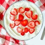 A glass bowl of whipped cream topped with sliced strawberries, styled like a Strawberry Rhubarb Trifle, sits on a red and white checkered cloth next to a spoon and plates.