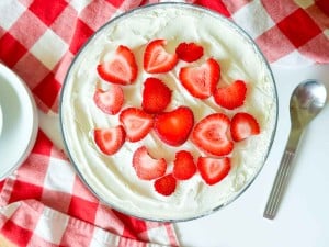 A glass bowl of whipped cream topped with sliced strawberries, styled like a Strawberry Rhubarb Trifle, sits on a red and white checkered cloth next to a spoon and plates.