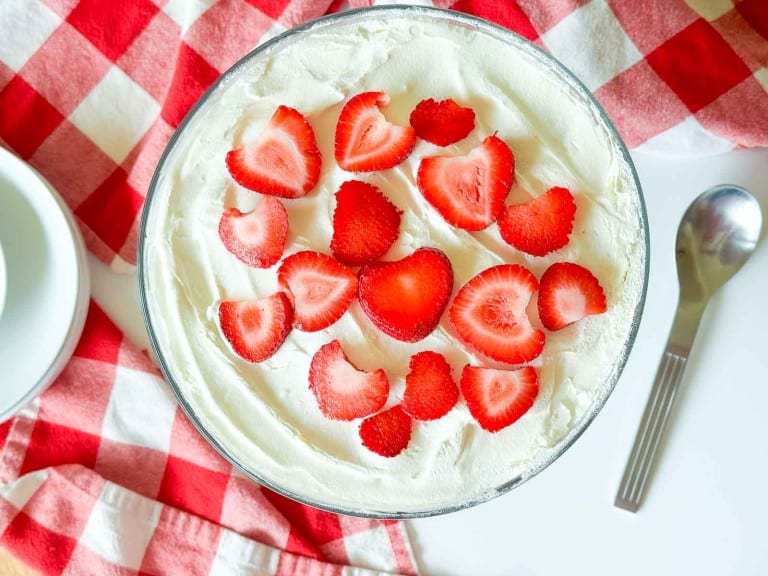 A glass bowl of whipped cream topped with sliced strawberries, styled like a Strawberry Rhubarb Trifle, sits on a red and white checkered cloth next to a spoon and plates.