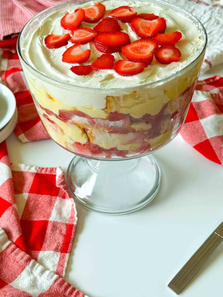 A Strawberry Rhubarb Trifle fills a glass dish with layers of cake, cream, and strawberries, sitting on a white table next to a red and white checkered cloth.