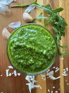 A glass bowl of green pesto sauce sits on a wooden surface, surrounded by arugula leaves, garlic cloves, and scattered cheese shavings.