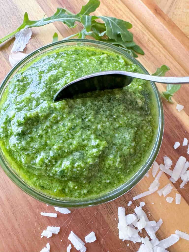 A glass bowl of green pesto with a spoon in it, placed on a wooden surface with arugula leaves and shaved Parmesan cheese nearby.