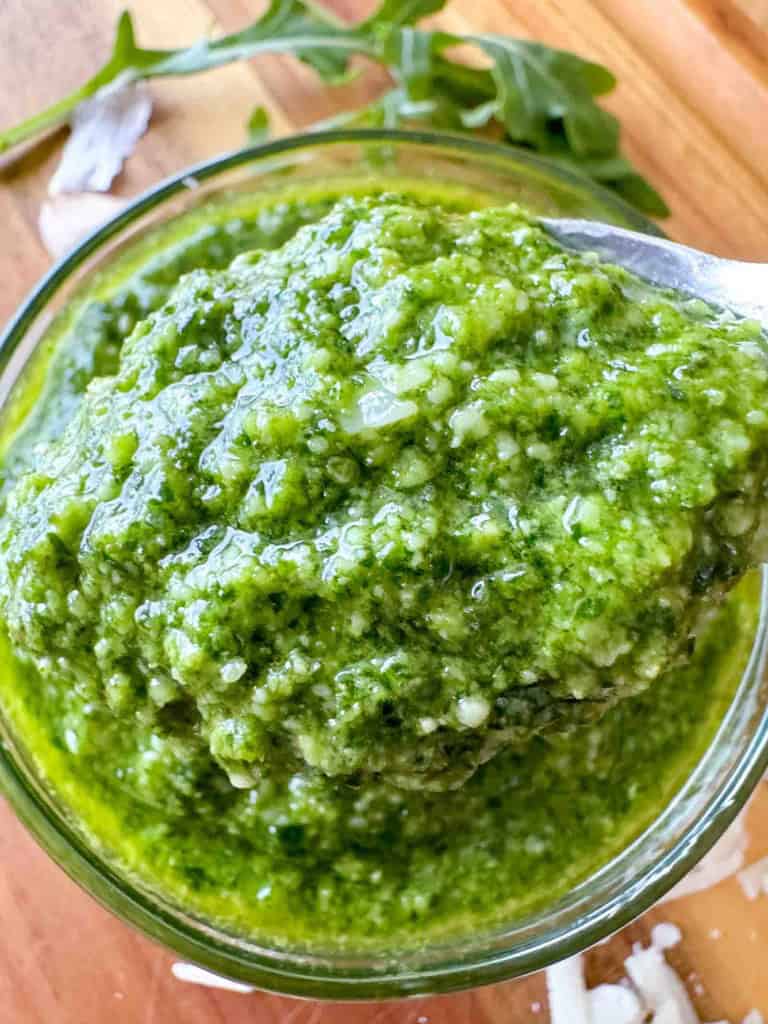Close-up of a spoon scooping green pesto sauce from a glass bowl, with fresh arugula leaves and shredded cheese in the background.