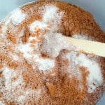 A mixing bowl filled with brown sugar and white granulated sugar being stirred with a wooden spoon.