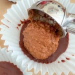 A metal scoop places a portion of brown filling onto chocolate in a white paper cupcake liner.