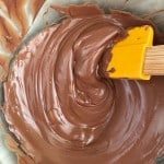 A mixing bowl with smooth, melted chocolate being stirred with a yellow spatula.