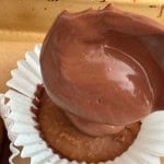 A spoon is spreading chocolate batter into a white paper cupcake liner on a baking tray.