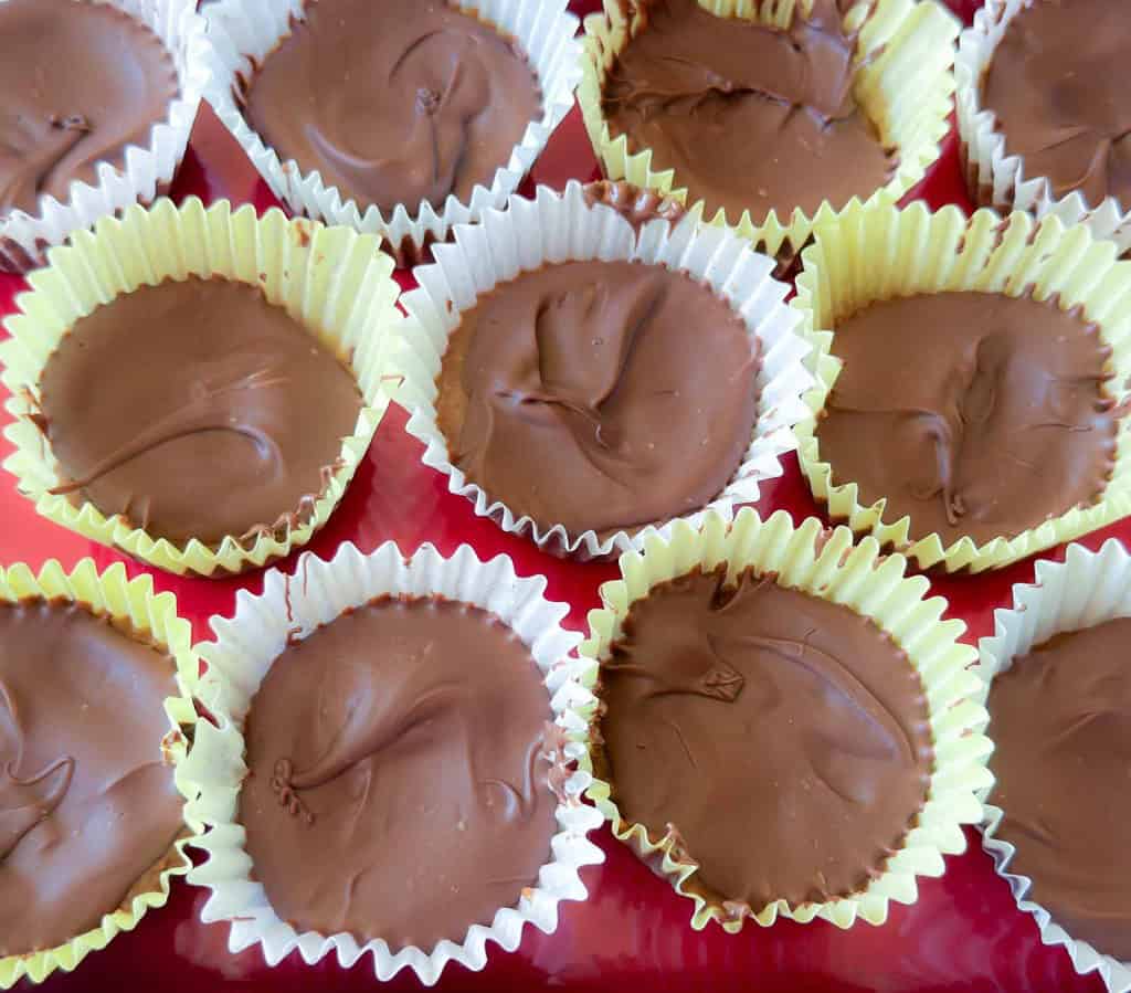 Rows of homemade chocolate cups in white paper liners are arranged closely together on a red surface.