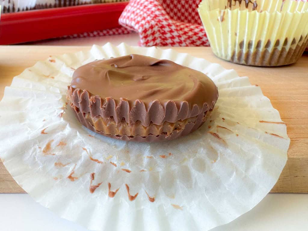 A chocolate peanut butter cup sits on an opened white paper liner, with another treat and a red checkered cloth in the background.
