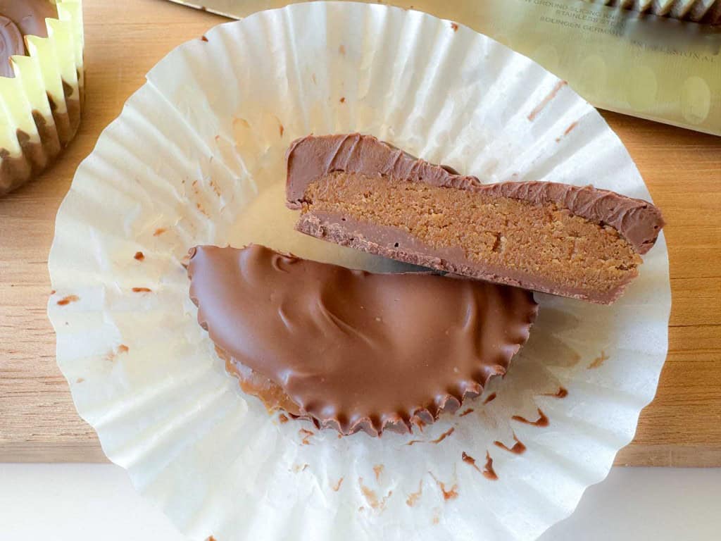 A chocolate peanut butter cup, partially cut in half to show the inside, sits on a white paper liner on a wooden surface.