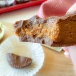A hand holding a half-eaten chocolate peanut butter cup, showing the peanut butter filling. Unwrapped chocolate and more cups are visible in the background.