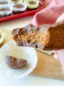 A hand holding a half-eaten chocolate peanut butter cup, showing the peanut butter filling. Unwrapped chocolate and more cups are visible in the background.