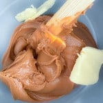 A wooden spoon mixing light brown peanut butter and chunks of solid butter in a bowl.