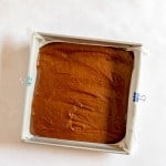 A square baking pan lined with parchment paper holds chocolate brownie batter, secured on two sides with binder clips, on a white surface.