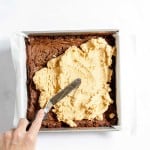 A hand spreading a layer of cookie dough over baked brownie batter in a parchment-lined square baking pan.