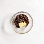 A glass bowl containing chocolate chips and a chunk of butter on a white surface.