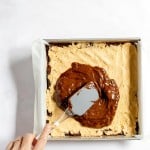 A hand spreads melted chocolate over a layer of peanut butter mixture in a parchment-lined square baking pan.