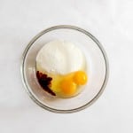 A glass bowl containing two eggs, granulated sugar, and a small amount of vanilla extract on a white surface.