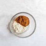 A glass bowl containing a mix of flour, cocoa powder, and a white powder, likely baking soda or powder, on a white surface.