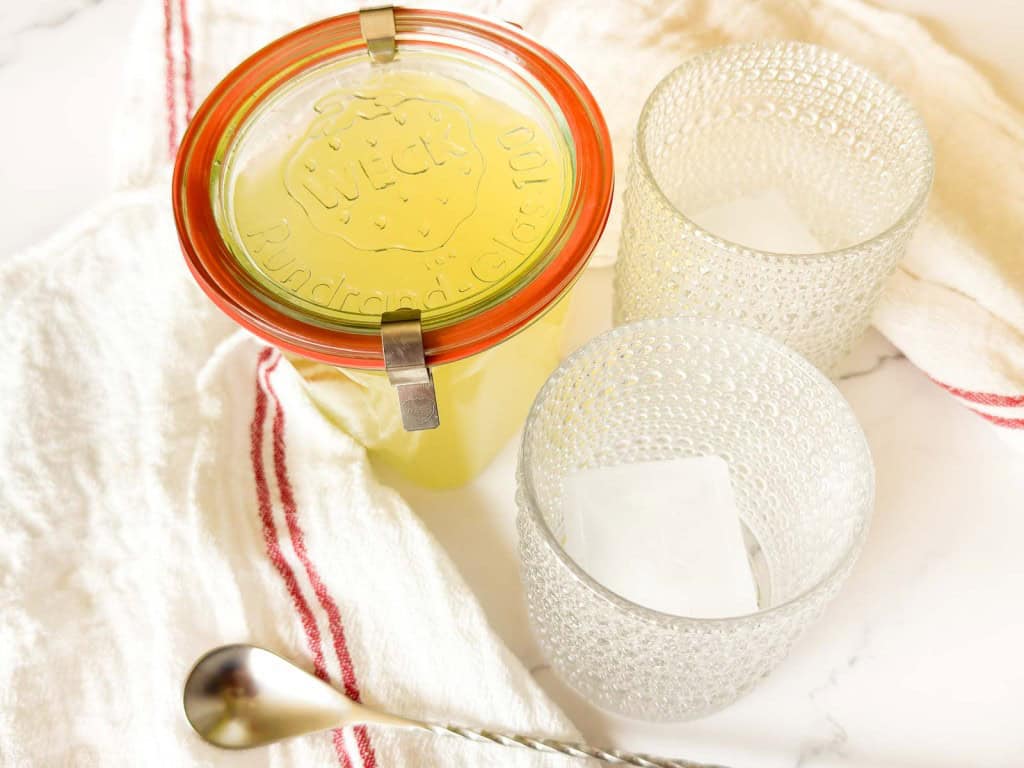 A glass jar of Classic Lemonade Syrup with a sealed lid sits beside two empty textured glasses with ice, a metal spoon, and a white cloth with red stripes.