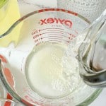 A glass measuring cup with Classic Lemonade Syrup being poured in, viewed from above, with measurement markings visible on the side.