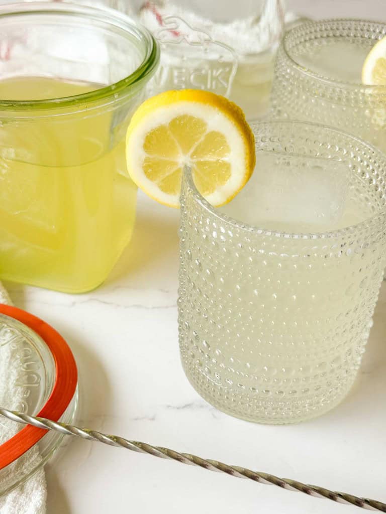 Two textured glasses of lemonade with ice and lemon slices, made with Classic Lemonade Syrup, sit next to a glass jar filled with more lemonade on a white surface.